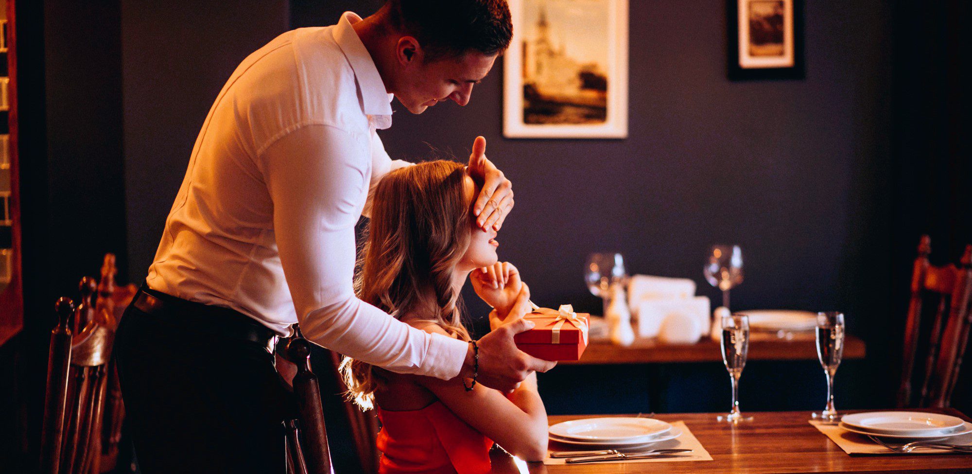 elegant couple in restaurant, her sitting at the table, him standing behind her, holding her eyes closed and offering her a present as surprise